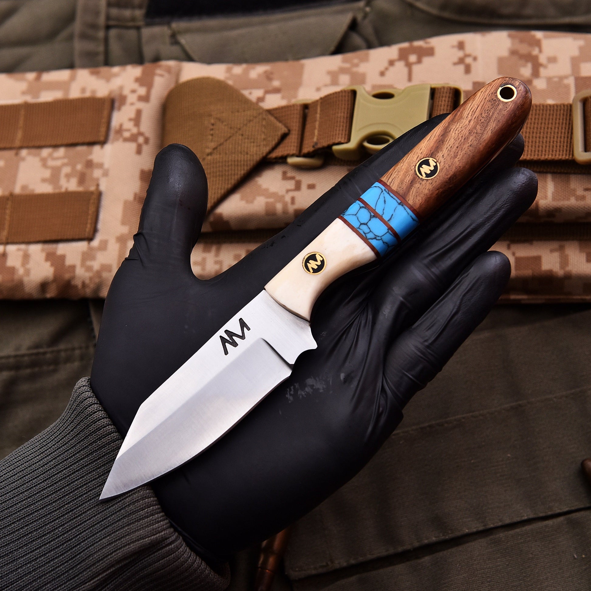 Knife with wooden handle and blue accent held by a gloved hand, with camouflage background