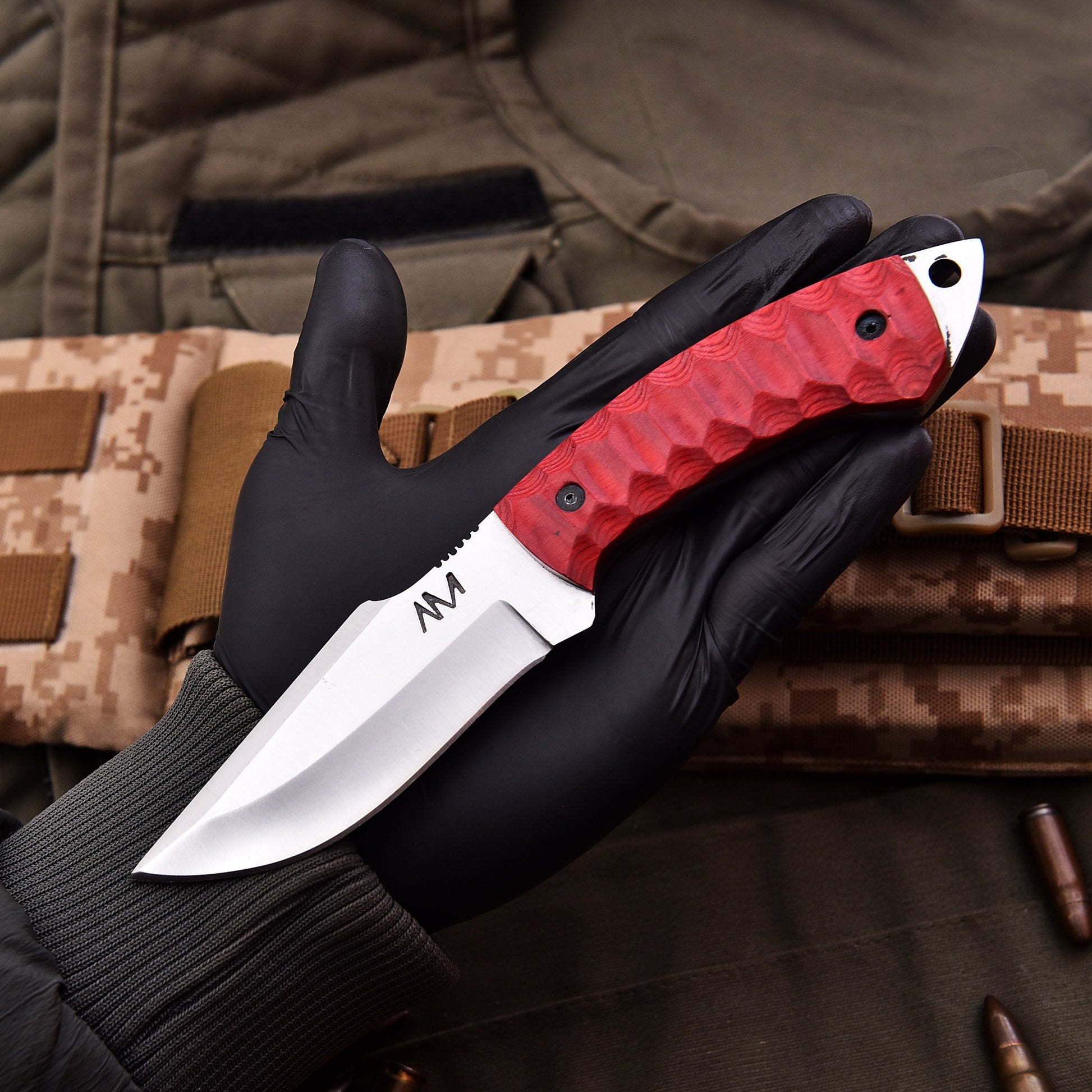 Knife with red handle held by a gloved hand against a camouflage background